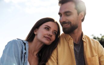 A woman with long brown hair leans her head affectionately on a man's shoulder. The man, with a beard and short brown hair, looks at her fondly. Both are outdoors in a sunlit setting with a clear sky in the background.