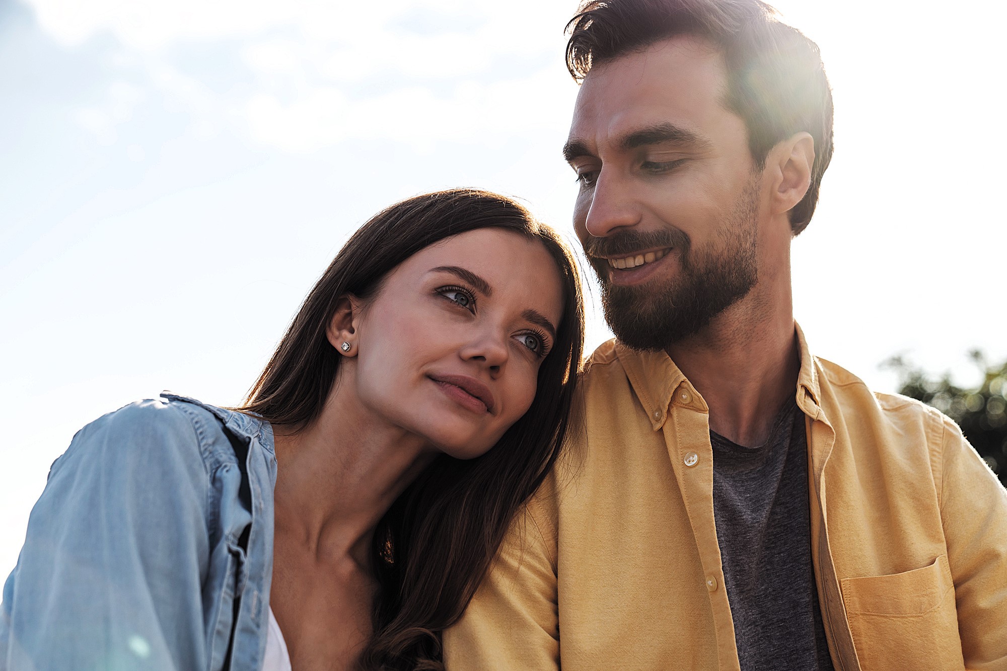 A woman with long brown hair leans her head affectionately on a man's shoulder. The man, with a beard and short brown hair, looks at her fondly. Both are outdoors in a sunlit setting with a clear sky in the background.