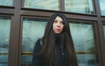 A woman with long dark hair is standing in front of a window with a wooden frame. She is wearing a black coat and a headband, gazing upwards with a thoughtful expression. The background shows reflections of buildings.