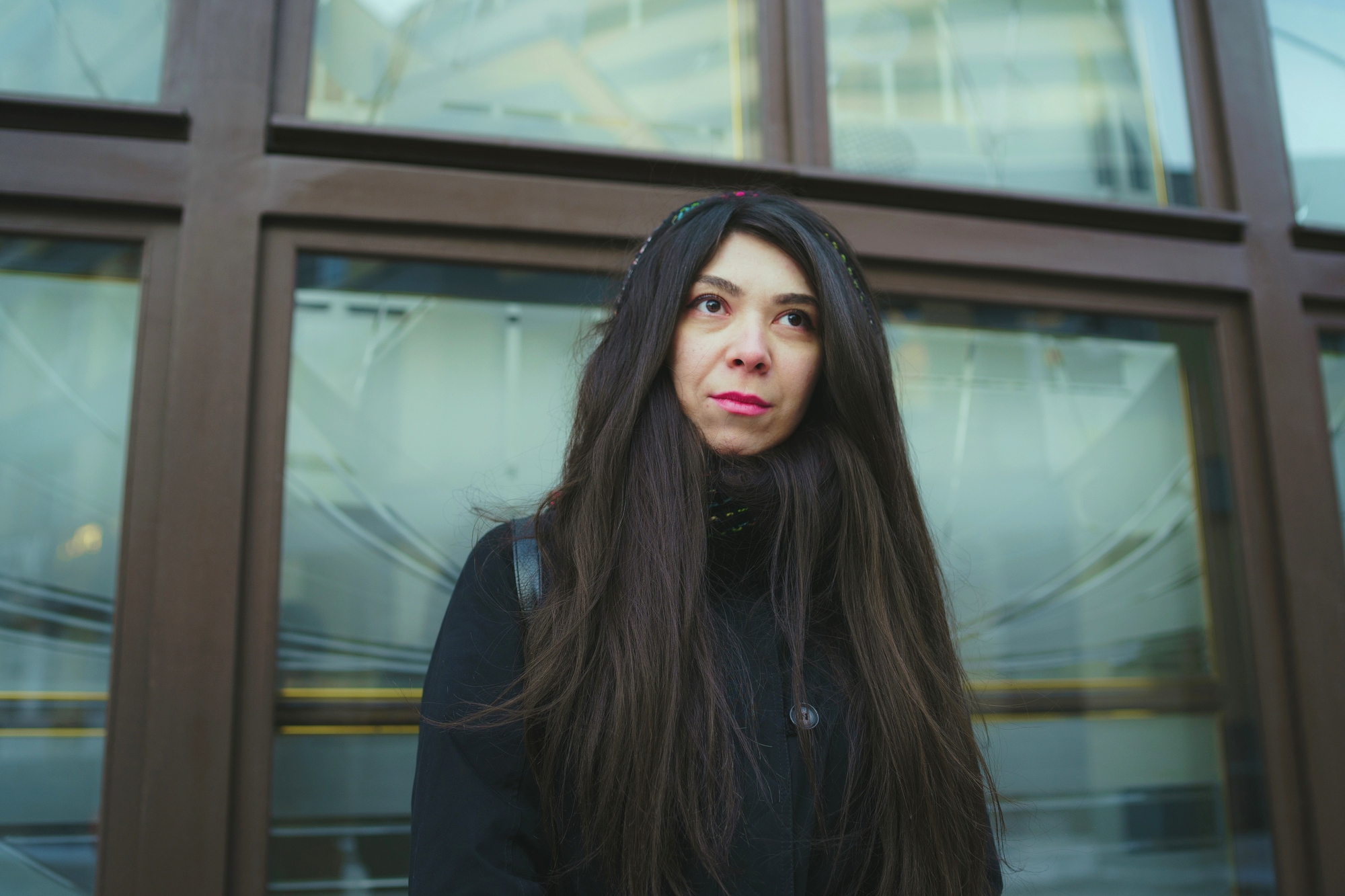 A woman with long dark hair is standing in front of a window with a wooden frame. She is wearing a black coat and a headband, gazing upwards with a thoughtful expression. The background shows reflections of buildings.