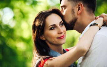 A woman with long dark hair smiles as a man with short hair kisses her forehead. They are outdoors with a blurred green leafy background, suggesting a park or garden setting. The woman is wearing a denim jacket.