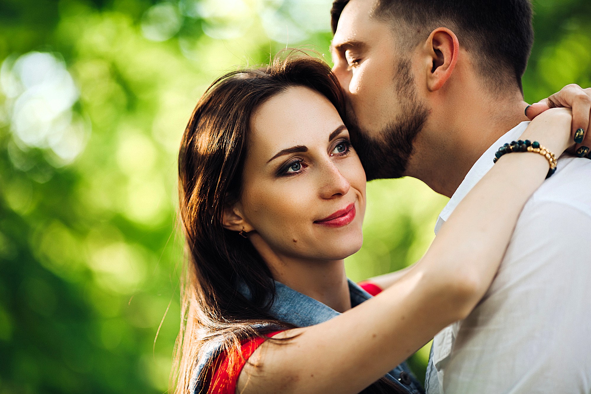 A woman with long dark hair smiles as a man with short hair kisses her forehead. They are outdoors with a blurred green leafy background, suggesting a park or garden setting. The woman is wearing a denim jacket.