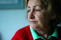 A pensive elderly woman with short brown hair is gazing out of the frame. She is wearing a red sweater over a green collared shirt. The background is softly blurred, creating a contemplative atmosphere.