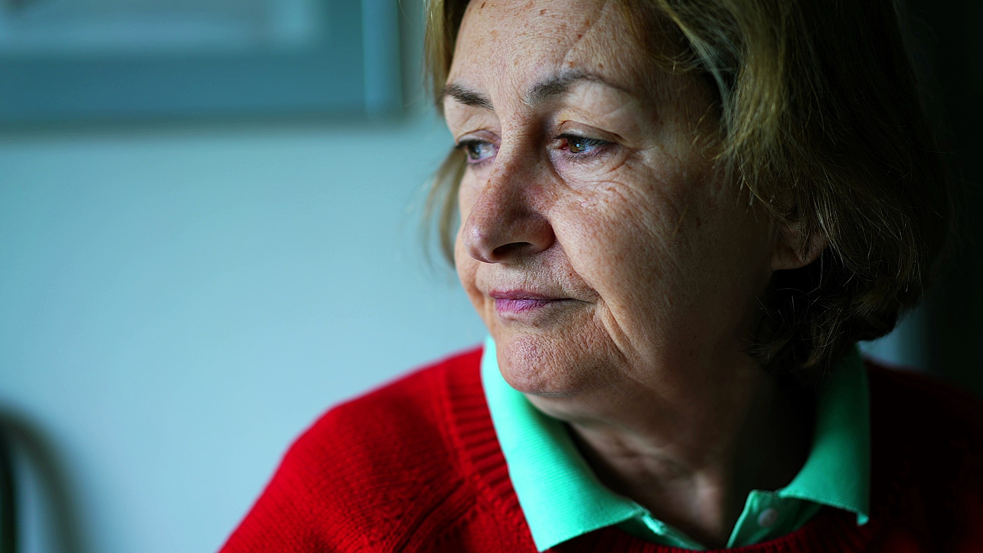 A pensive elderly woman with short brown hair is gazing out of the frame. She is wearing a red sweater over a green collared shirt. The background is softly blurred, creating a contemplative atmosphere.