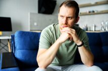 A man with short hair and a beard sits on a blue couch, looking contemplative. He rests his chin on his hands, wearing a light green shirt and a smartwatch, with a blurred office space in the background.