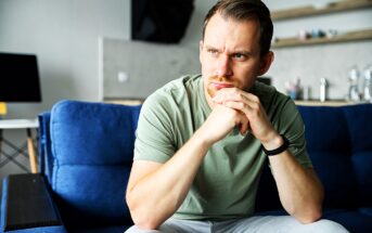 A man with short hair and a beard sits on a blue couch, looking contemplative. He rests his chin on his hands, wearing a light green shirt and a smartwatch, with a blurred office space in the background.