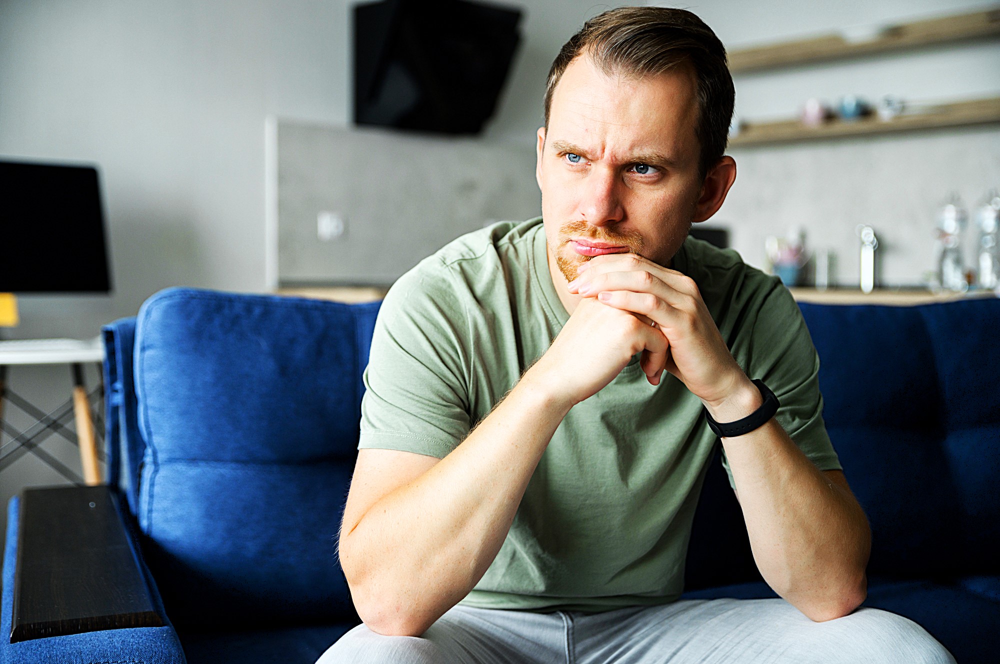 A man with short hair and a beard sits on a blue couch, looking contemplative. He rests his chin on his hands, wearing a light green shirt and a smartwatch, with a blurred office space in the background.