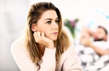 A thoughtful woman with long blonde hair rests her chin on her hand, looking to the side. She wears a light sweater. In the background, a person lies on a bed or sofa, blurred for depth.