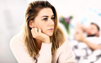 A thoughtful woman with long blonde hair rests her chin on her hand, looking to the side. She wears a light sweater. In the background, a person lies on a bed or sofa, blurred for depth.