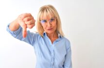 An older woman with blonde hair and glasses stands against a plain white background, looking directly at the camera. She is wearing a light blue shirt and giving a thumbs-down gesture with her right hand.