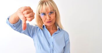 An older woman with blonde hair and glasses stands against a plain white background, looking directly at the camera. She is wearing a light blue shirt and giving a thumbs-down gesture with her right hand.