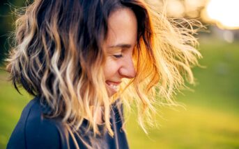 A woman with shoulder-length wavy hair smiles while looking down. Her hair is highlighted by the sunlight, creating a warm glow. She is outdoors, with a blurred green background.