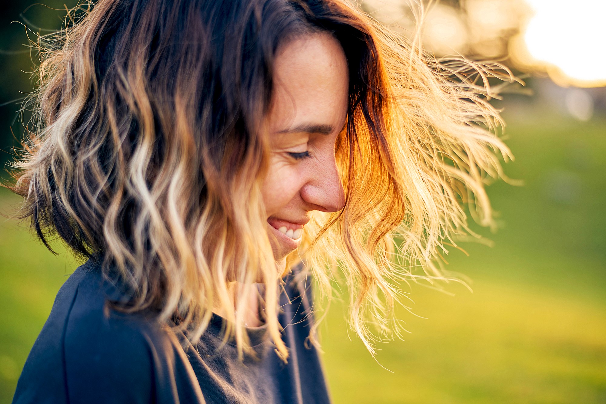 A woman with shoulder-length wavy hair smiles while looking down. Her hair is highlighted by the sunlight, creating a warm glow. She is outdoors, with a blurred green background.