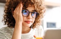 A person with curly hair and glasses is looking at a laptop screen, holding a glass of orange juice. They appear thoughtful or focused, with one hand resting on their face. They're wearing a striped shirt and earphones, in a softly lit room.