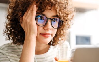 A person with curly hair and glasses is looking at a laptop screen, holding a glass of orange juice. They appear thoughtful or focused, with one hand resting on their face. They're wearing a striped shirt and earphones, in a softly lit room.