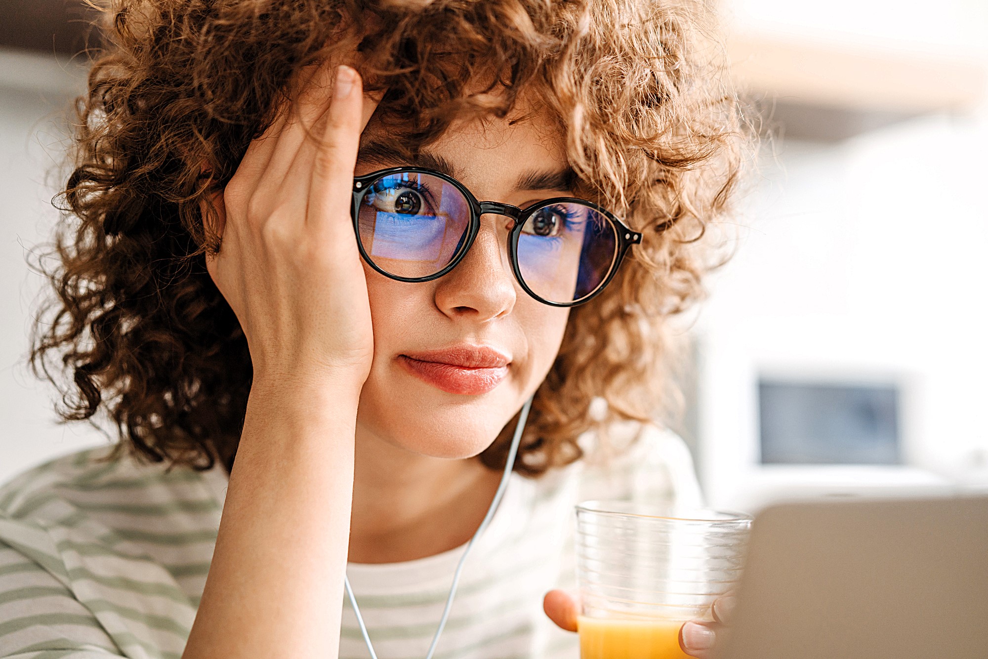 A person with curly hair and glasses is looking at a laptop screen, holding a glass of orange juice. They appear thoughtful or focused, with one hand resting on their face. They're wearing a striped shirt and earphones, in a softly lit room.