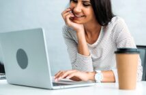 A woman in a striped shirt is sitting at a desk, smiling while looking at her laptop. Her hand rests on her chin. A paper coffee cup is placed on the desk beside her.