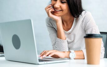 A woman in a striped shirt is sitting at a desk, smiling while looking at her laptop. Her hand rests on her chin. A paper coffee cup is placed on the desk beside her.