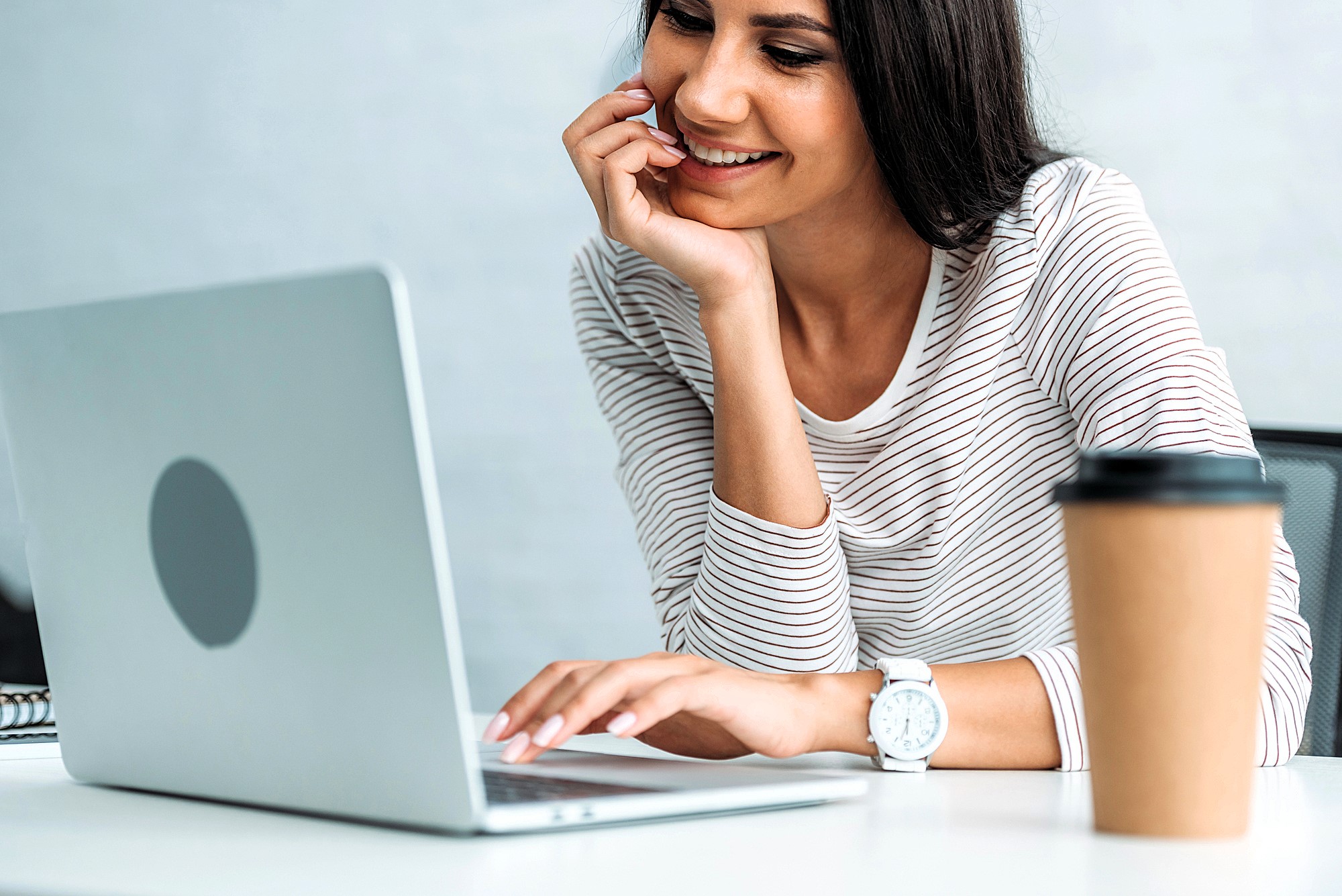 A woman in a striped shirt is sitting at a desk, smiling while looking at her laptop. Her hand rests on her chin. A paper coffee cup is placed on the desk beside her.