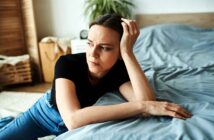 A woman in a black shirt and jeans leans on a bed with a pensive expression. She rests her head on her hand, while gazing thoughtfully. A plant and wicker basket are visible in the softly lit background.