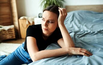 A woman in a black shirt and jeans leans on a bed with a pensive expression. She rests her head on her hand, while gazing thoughtfully. A plant and wicker basket are visible in the softly lit background.