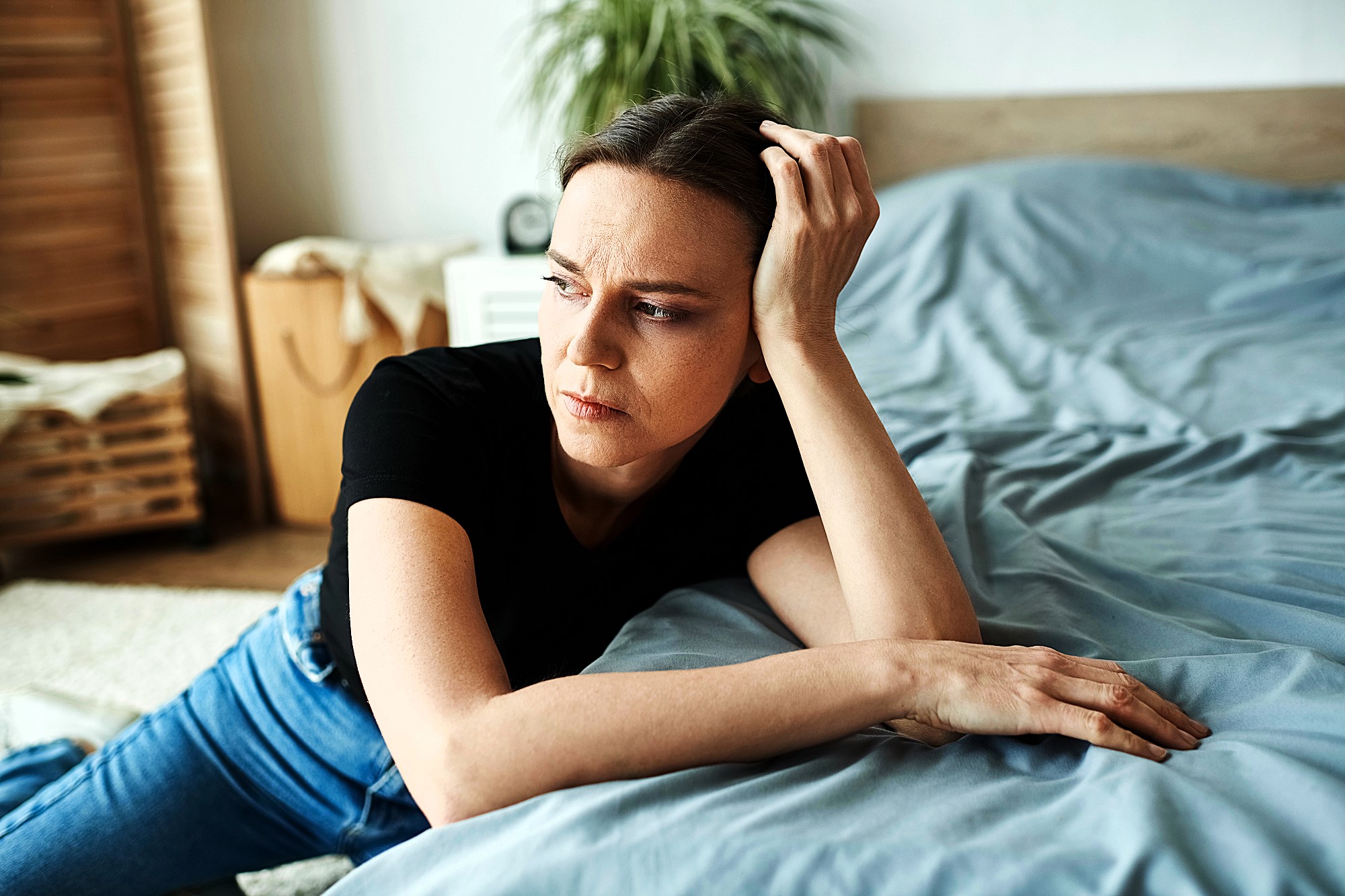 A woman in a black shirt and jeans leans on a bed with a pensive expression. She rests her head on her hand, while gazing thoughtfully. A plant and wicker basket are visible in the softly lit background.