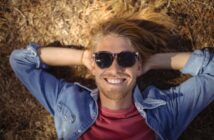 A person with long blonde hair and sunglasses is lying on the ground, smiling. They are wearing a denim shirt over a red t-shirt. The sun casts their shadow on dry grass and leaves.