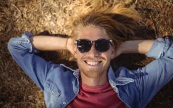 A person with long blonde hair and sunglasses is lying on the ground, smiling. They are wearing a denim shirt over a red t-shirt. The sun casts their shadow on dry grass and leaves.