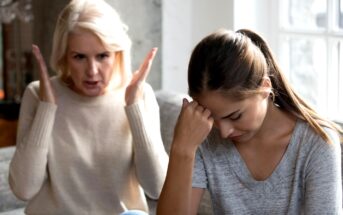 An older woman with gray hair appears frustrated, gesturing with her hands while sitting on a couch. A younger woman next to her, with brown hair tied back, looks upset with her head down, resting her hand on her forehead.