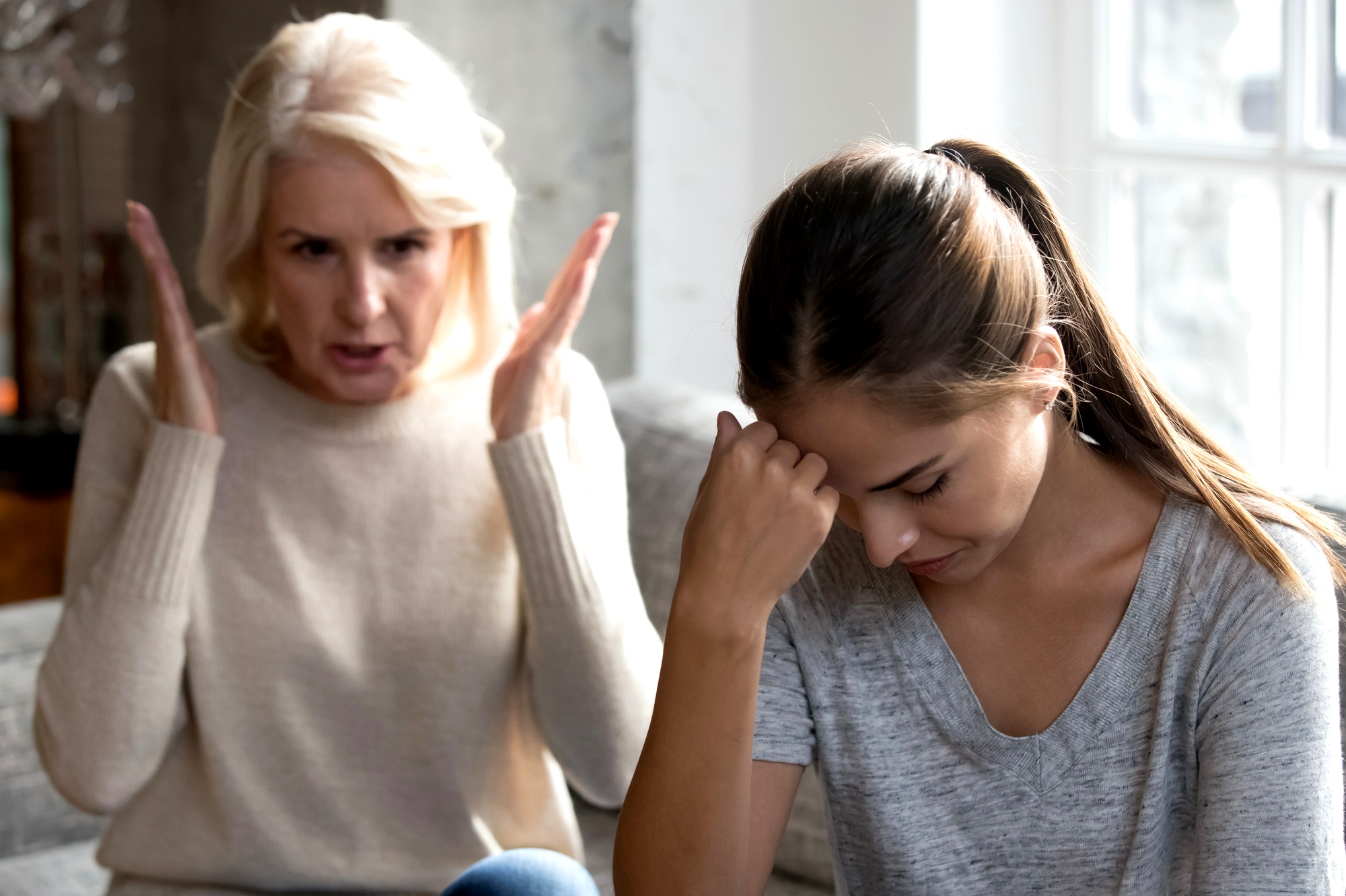 An older woman with gray hair appears frustrated, gesturing with her hands while sitting on a couch. A younger woman next to her, with brown hair tied back, looks upset with her head down, resting her hand on her forehead.