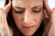 A woman is pressing her temples with her fingers, eyes closed, appearing to be in pain or discomfort, possibly experiencing a headache. Her expression is tense, with a slight frown.