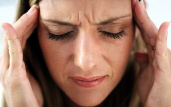 A woman is pressing her temples with her fingers, eyes closed, appearing to be in pain or discomfort, possibly experiencing a headache. Her expression is tense, with a slight frown.