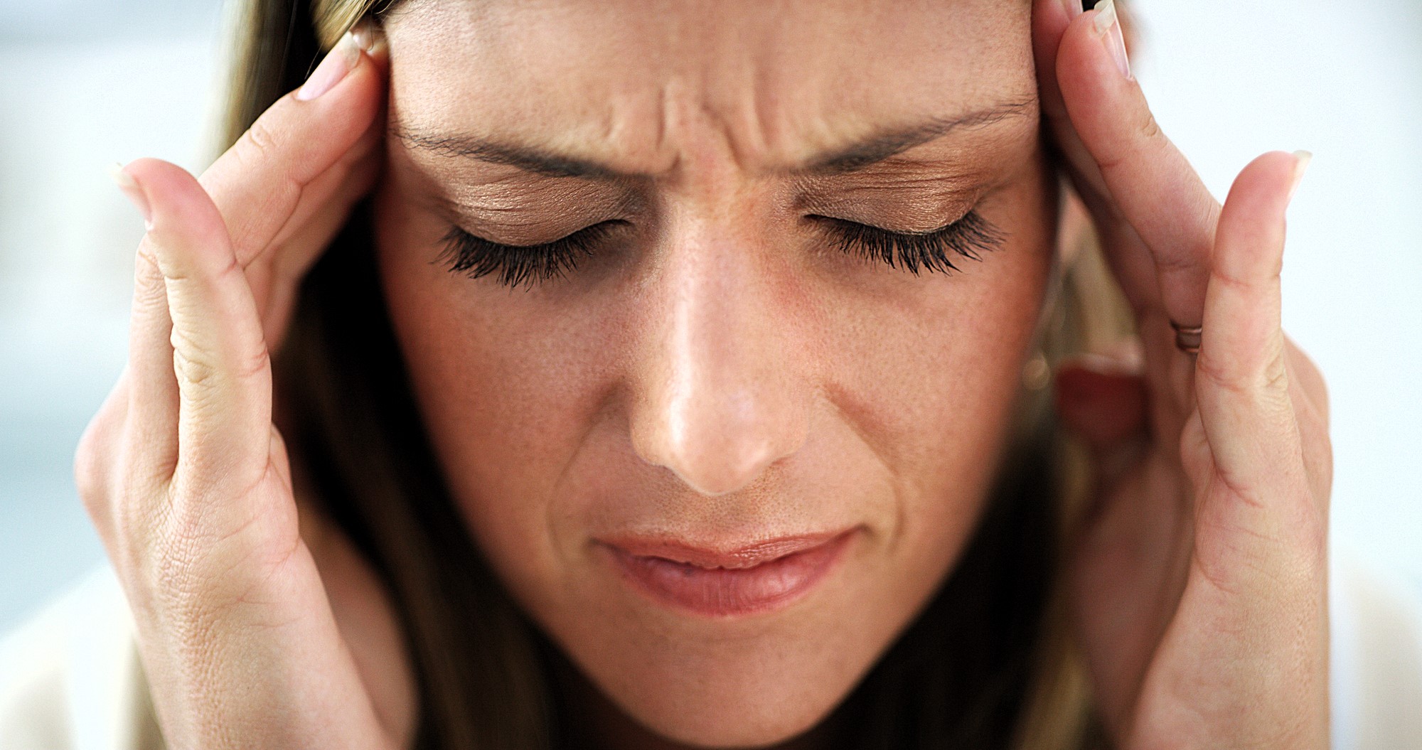 A woman is pressing her temples with her fingers, eyes closed, appearing to be in pain or discomfort, possibly experiencing a headache. Her expression is tense, with a slight frown.