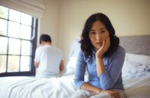 A woman sits on a bed looking contemplative, with her head resting on her hand. In the background, a man sits facing away, near a window with crossed arms. The room is softly lit by natural light.