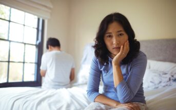 A woman sits on a bed looking contemplative, with her head resting on her hand. In the background, a man sits facing away, near a window with crossed arms. The room is softly lit by natural light.