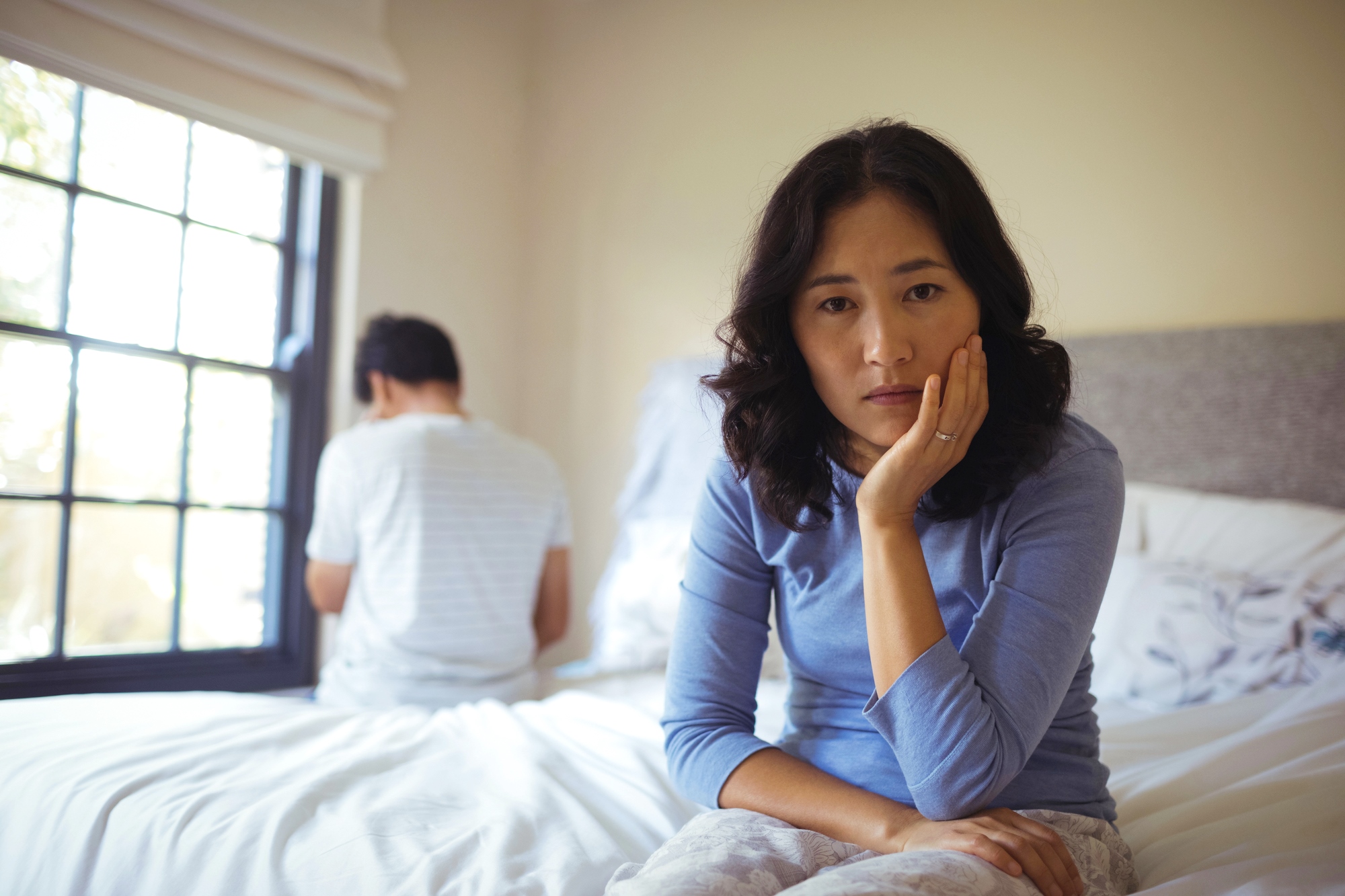 A woman sits on a bed looking contemplative, with her head resting on her hand. In the background, a man sits facing away, near a window with crossed arms. The room is softly lit by natural light.