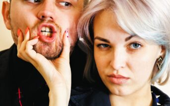 A woman with short hair playfully squeezes a man's face, making his lips pucker humorously. Both have expressions of amusement and skepticism. The man's beard is trimmed, and they are in close proximity, suggesting a lighthearted moment.