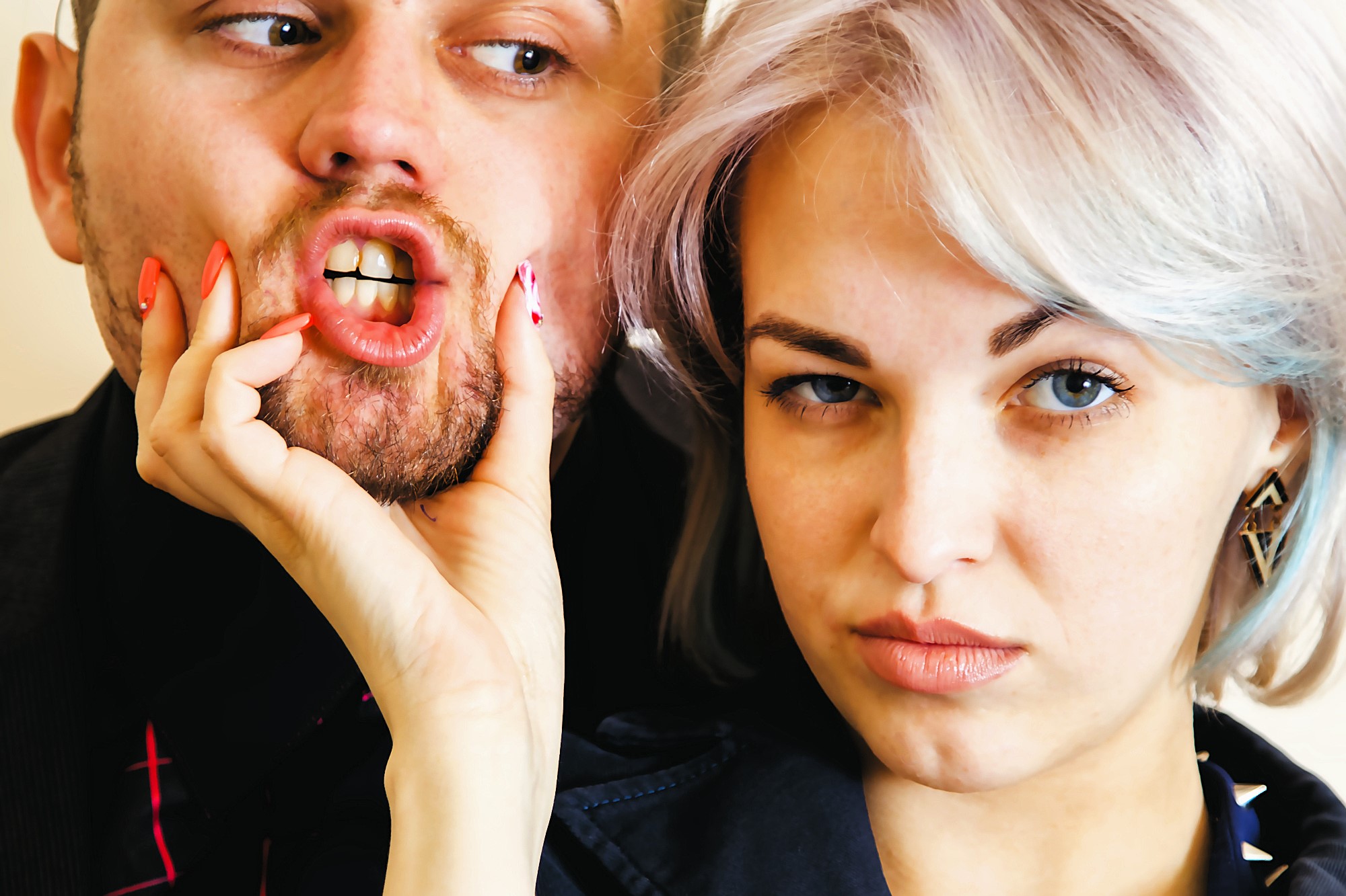 A woman with short hair playfully squeezes a man's face, making his lips pucker humorously. Both have expressions of amusement and skepticism. The man's beard is trimmed, and they are in close proximity, suggesting a lighthearted moment.