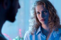 A woman with curly hair and a pensive expression looks at a blurred man in the foreground. The scene is softly lit with blue and pink tones, suggesting a contemplative atmosphere.