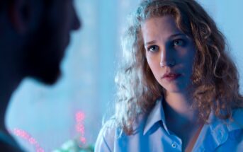 A woman with curly hair and a pensive expression looks at a blurred man in the foreground. The scene is softly lit with blue and pink tones, suggesting a contemplative atmosphere.