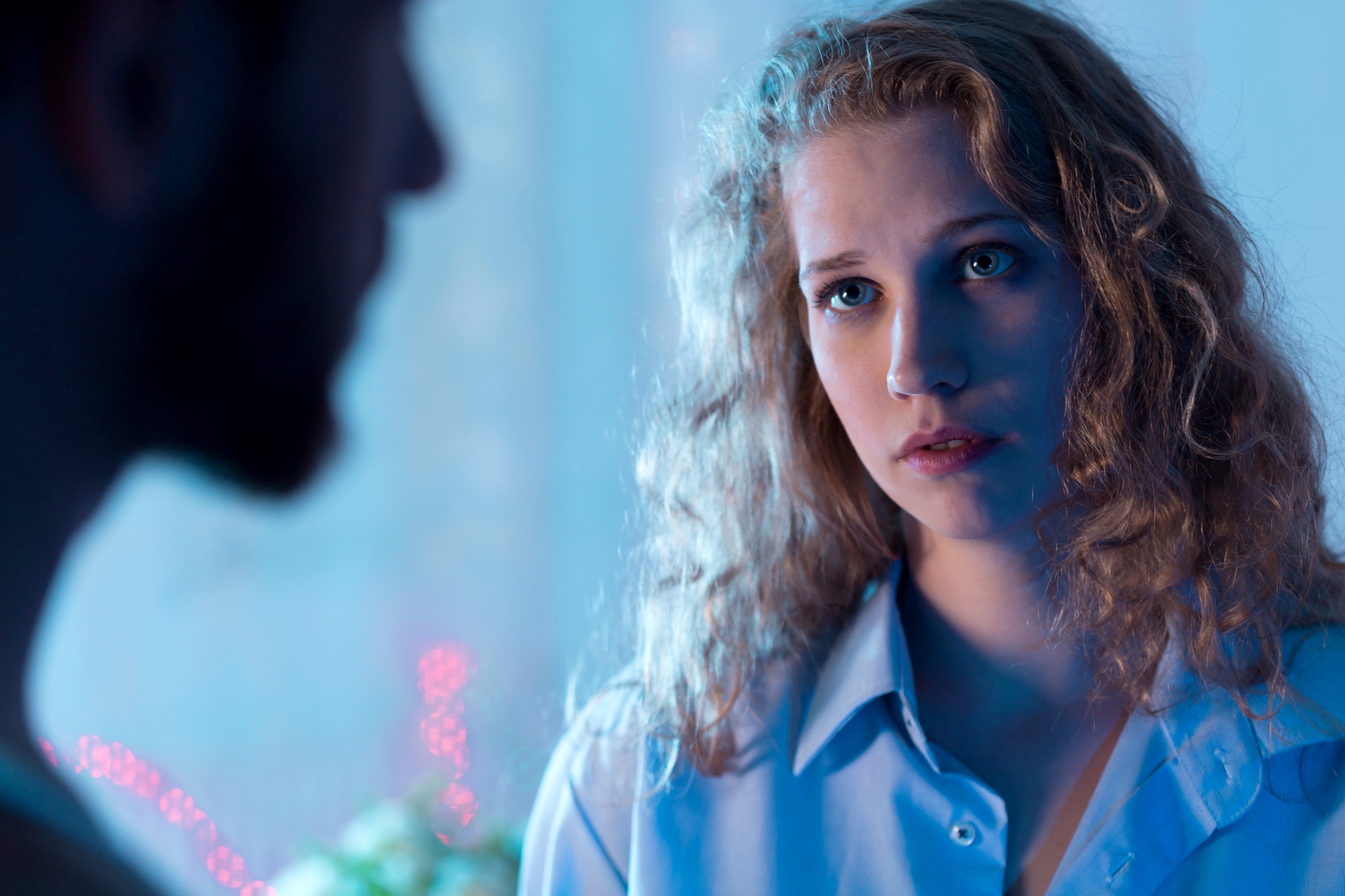 A woman with curly hair and a pensive expression looks at a blurred man in the foreground. The scene is softly lit with blue and pink tones, suggesting a contemplative atmosphere.