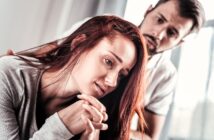 A woman with long red hair looks thoughtful and troubled, resting her head on her hands. A man with a concerned expression stands behind her, gently placing a hand on her shoulder. They are indoors with soft lighting.