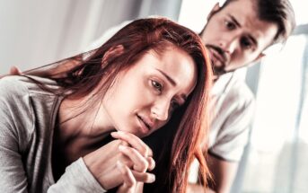 A woman with long red hair looks thoughtful and troubled, resting her head on her hands. A man with a concerned expression stands behind her, gently placing a hand on her shoulder. They are indoors with soft lighting.