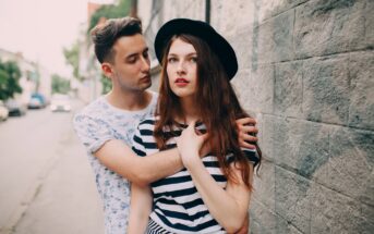 A young man and woman stand close together by a stone wall. The man, wearing a patterned shirt, has his arm around the woman, who is dressed in a striped top and a black hat. They both have a serious expression.