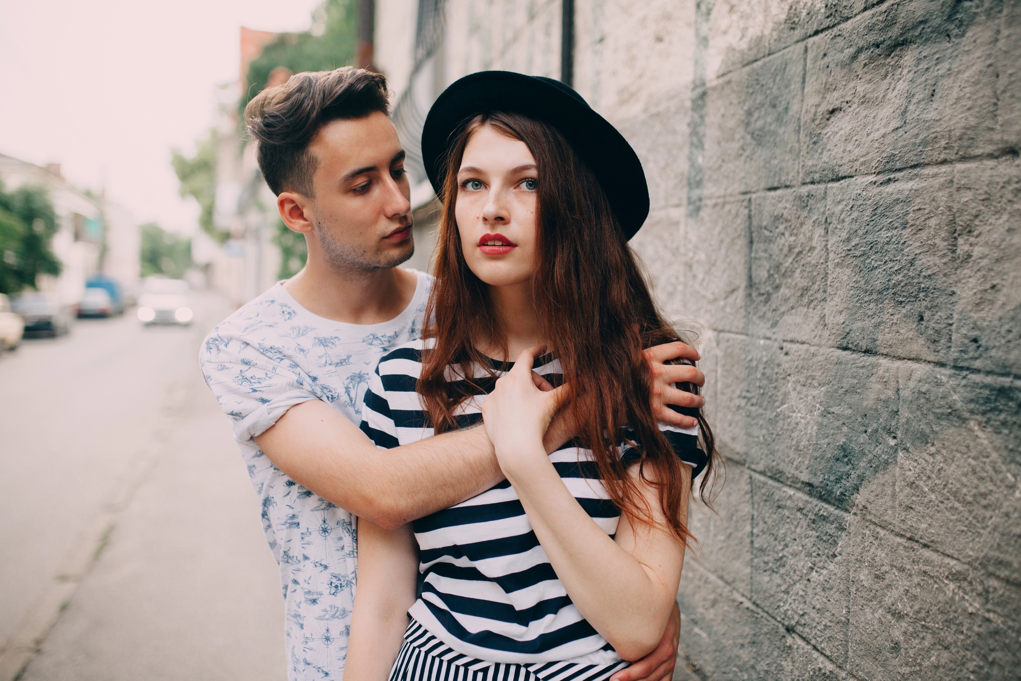 A young man and woman stand close together by a stone wall. The man, wearing a patterned shirt, has his arm around the woman, who is dressed in a striped top and a black hat. They both have a serious expression.