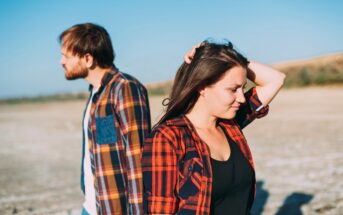 A man and woman in casual plaid shirts stand back-to-back outdoors, with the woman looking down and touching her hair. The background shows an open, sunny landscape under a clear blue sky.