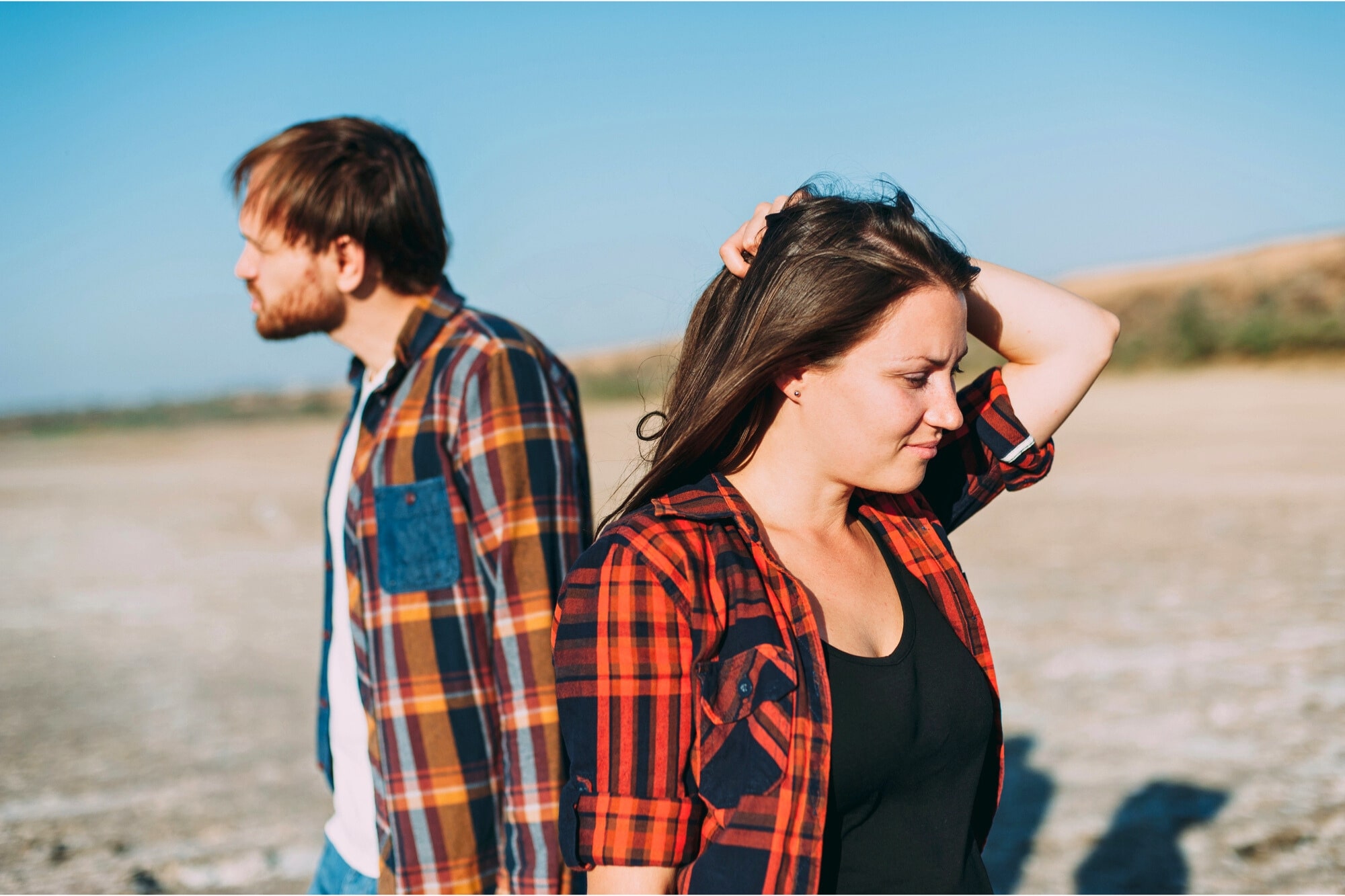 A man and woman in casual plaid shirts stand back-to-back outdoors, with the woman looking down and touching her hair. The background shows an open, sunny landscape under a clear blue sky.