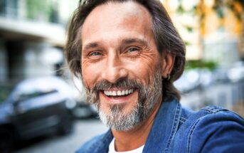 A middle-aged man with a beard is smiling warmly at the camera. He is wearing a denim jacket, and the background is a blurred urban setting with hints of parked cars and buildings.