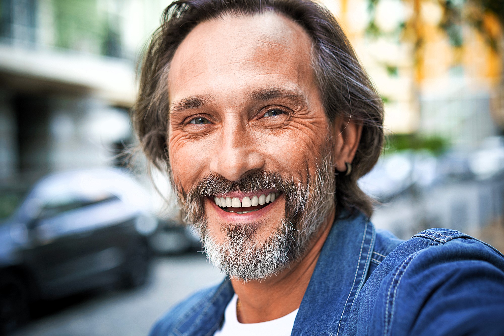 A middle-aged man with a beard is smiling warmly at the camera. He is wearing a denim jacket, and the background is a blurred urban setting with hints of parked cars and buildings.