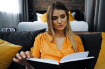 A woman with long hair is sitting on a couch, wearing an orange blouse, and reading a book. Behind her is a bed and yellow cushions. The room has a cozy, softly lit atmosphere.